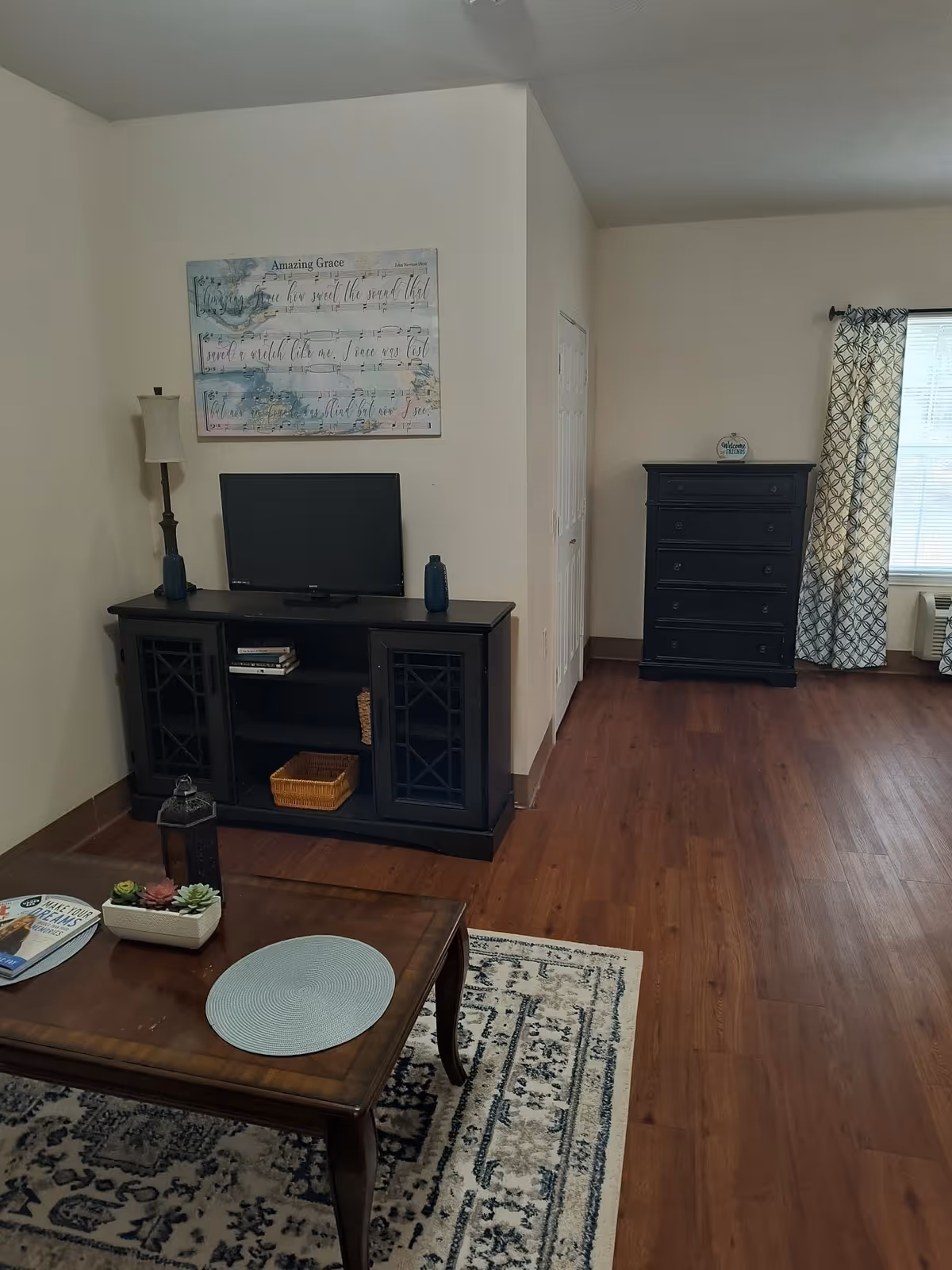 A living room area with a wooden coffee table on a patterned rug, a TV on a black cabinet with decorative items, a tall black dresser near a window with patterned curtains, and wooden flooring throughout.