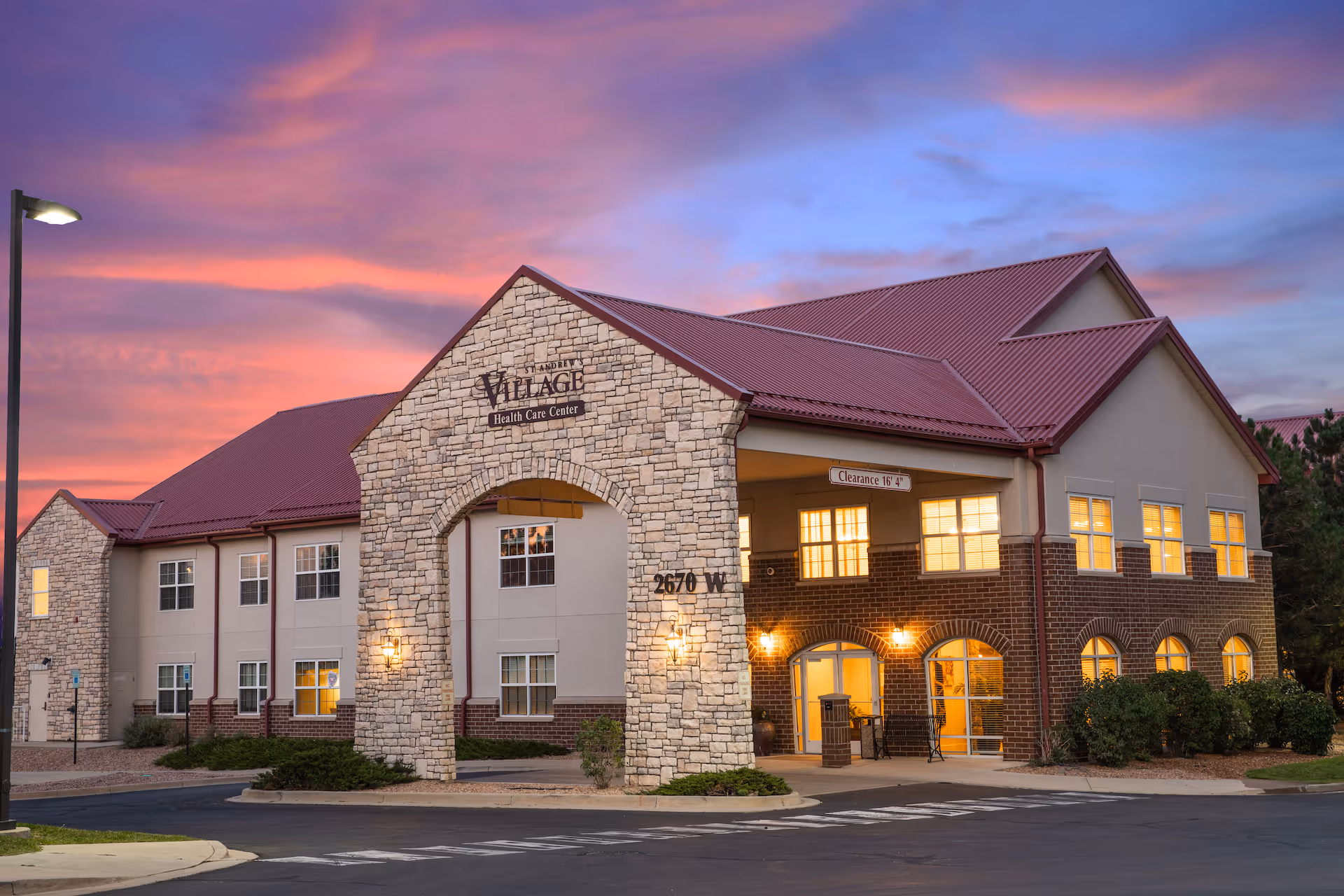 Exterior view of The Springs at St Andrew's Village Health Care Center building at dusk, featuring a stone and brick facade with warm interior lights glowing through the windows and a colorful sunset sky in the background.