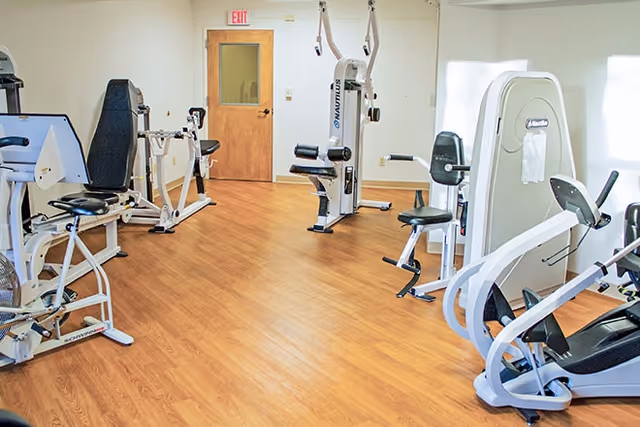 A fitness room with various exercise machines including stationary bikes and weight machines on a wooden floor. There is a wooden door with a window and an exit sign above it in the background.