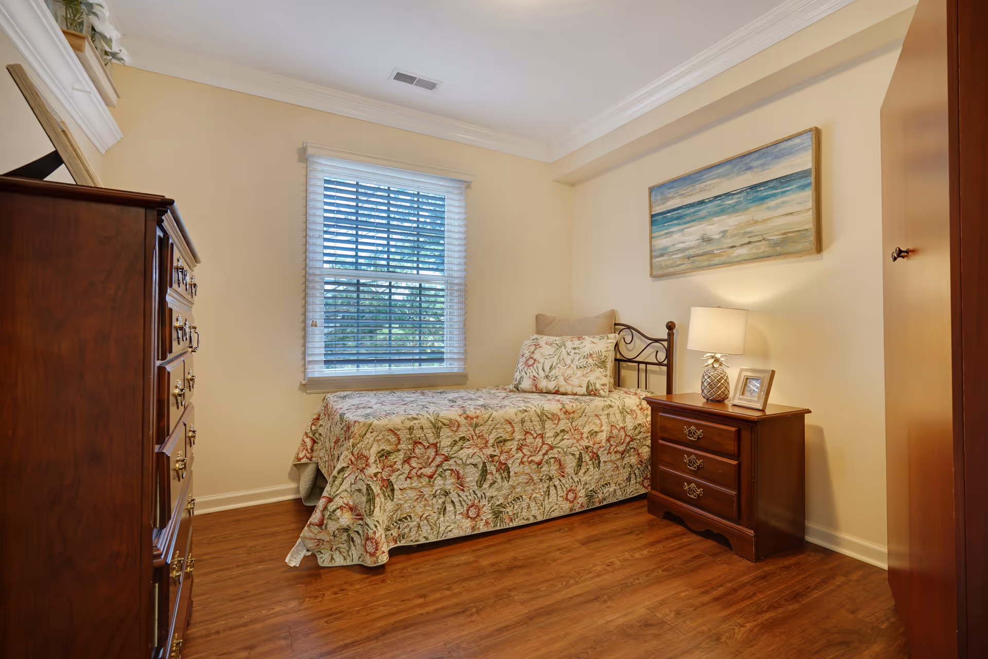 A cozy bedroom with a single bed covered in a floral patterned bedspread. There is a wooden nightstand with a lamp and a framed photo, a wooden dresser, and a window with blinds letting in natural light. A painting of a beach scene hangs on the wall above the bed.