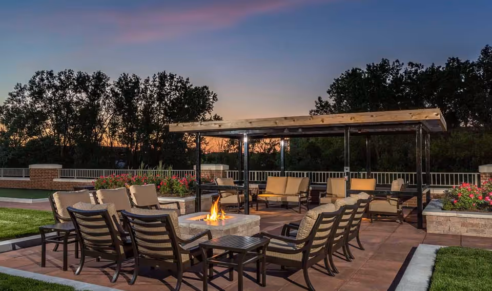 Outdoor patio area at sunset with cushioned chairs arranged around a fire pit, a covered seating area with sofas and chairs, surrounded by greenery and flowers.