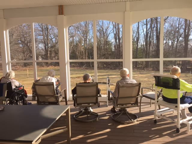Five elderly individuals sitting in chairs and a wheelchair on a screened porch, facing a yard with leafless trees under a clear sky.