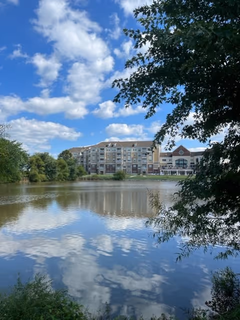 Multi-story residential building viewed across a calm pond, framed by trees under a blue sky with clouds.