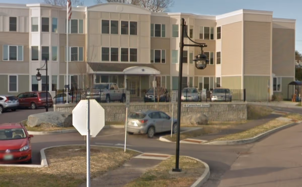 Front exterior of a three-story senior living building with parked cars, a flagpole, and a driveway.