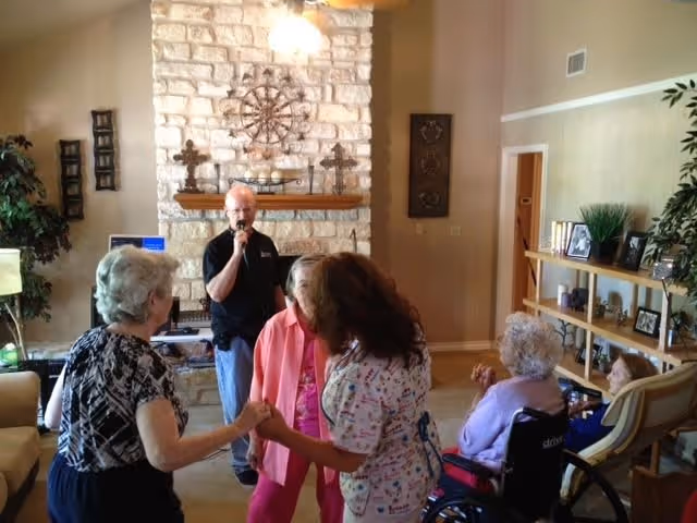 A group of elderly people and a caregiver are gathered in a living room with a stone fireplace. One man is holding a microphone, possibly singing or speaking, while two women hold hands and dance. Two other elderly women are seated, one in a wheelchair, watching the activity. The room is warmly decorated with plants, shelves with framed photos, and wall art.