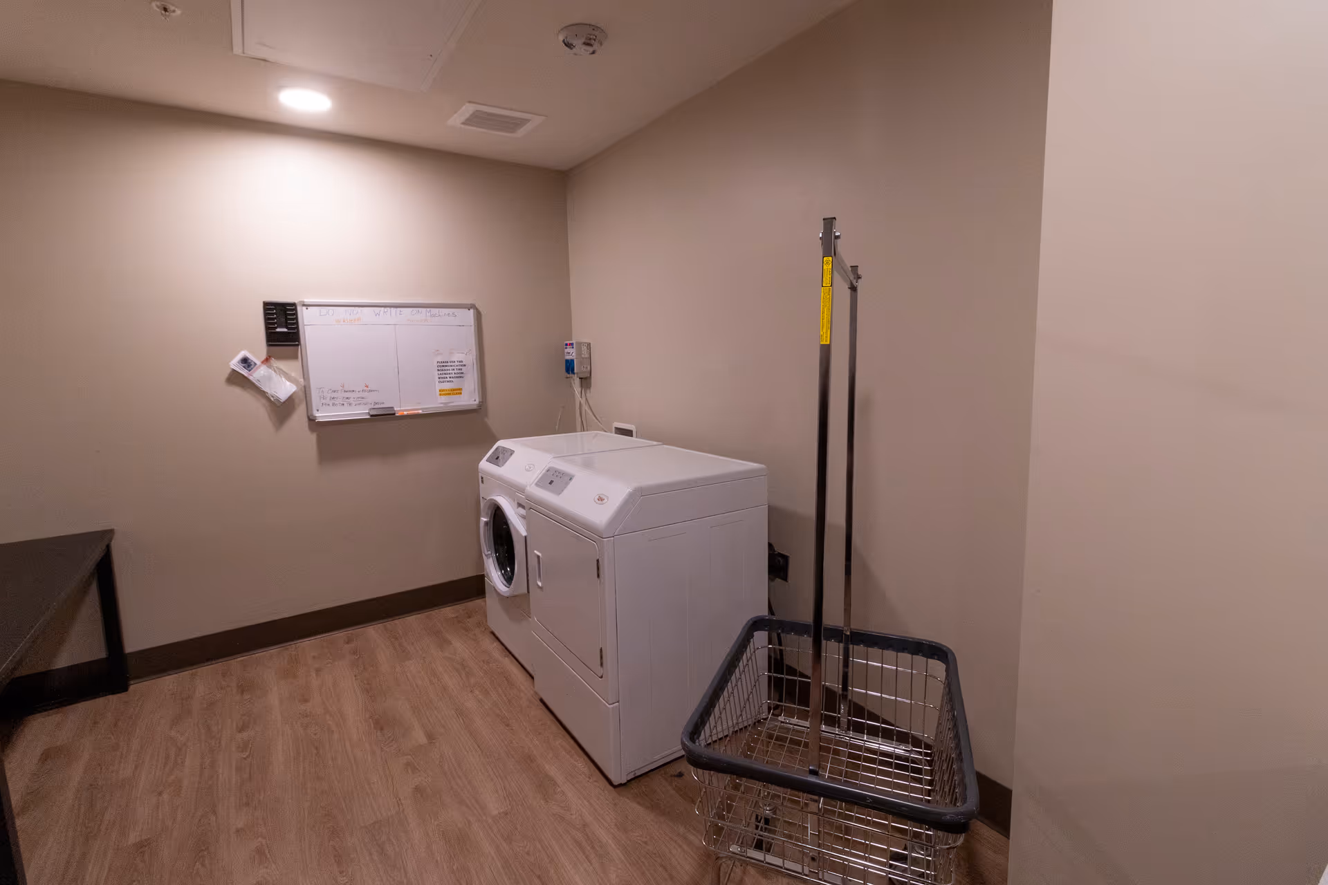 A laundry room with a washing machine and dryer side by side against a beige wall. There is a metal laundry cart with a handle in the foreground on the right. A whiteboard with notes and instructions is mounted on the wall above the machines. The floor is wood-patterned, and the room is lit by a ceiling light.
