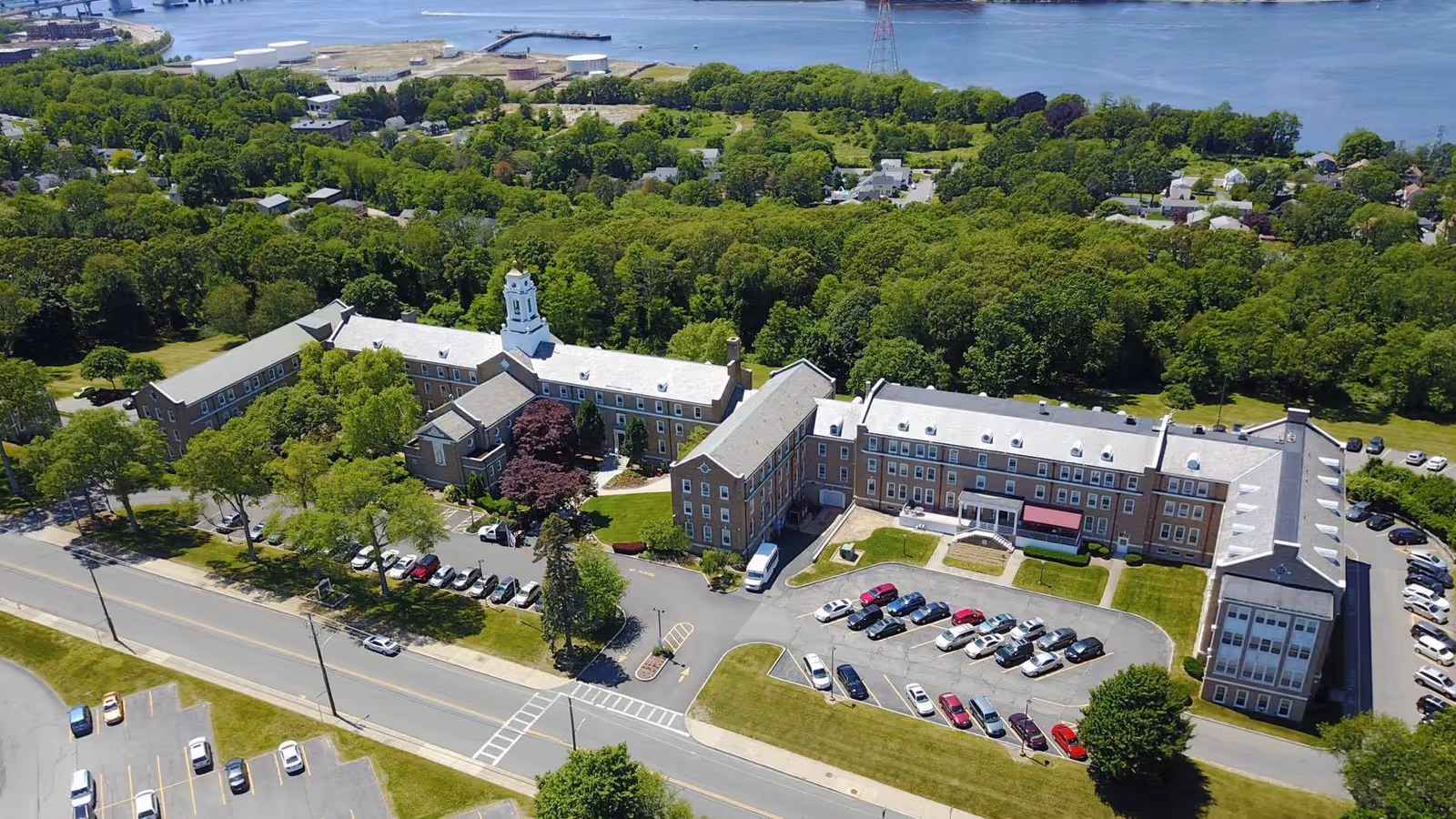 Aerial view of a large brick skilled nursing facility with a parking lot, landscaped grounds, surrounding trees, and a nearby waterfront.