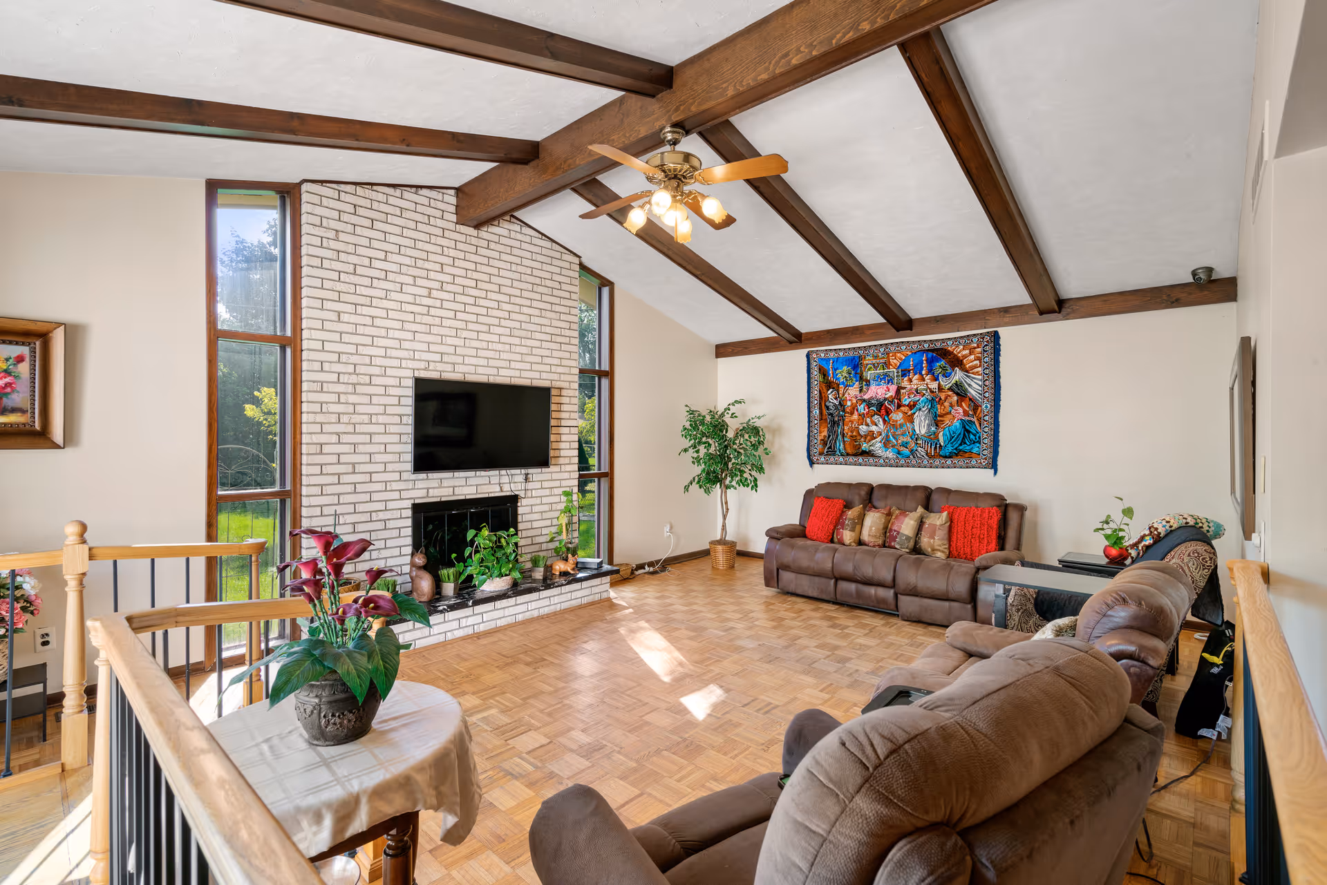 A spacious living room with wooden ceiling beams and a ceiling fan. The room features a white brick fireplace with a mounted flat-screen TV above it, flanked by tall narrow windows. There are two brown reclining sofas with colorful cushions, a small table with a potted plant, and a large decorative tapestry hanging on the wall behind one sofa. The floor is parquet wood, and there are additional plants and decorative items around the room.