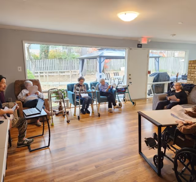 A group of elderly individuals seated in a common room with large windows showing an outdoor fenced area with a gazebo. Some people are sitting in chairs and one person is in a wheelchair, all holding papers. A man is playing a guitar near a laptop on a small table. The room has wooden flooring and light-colored walls.