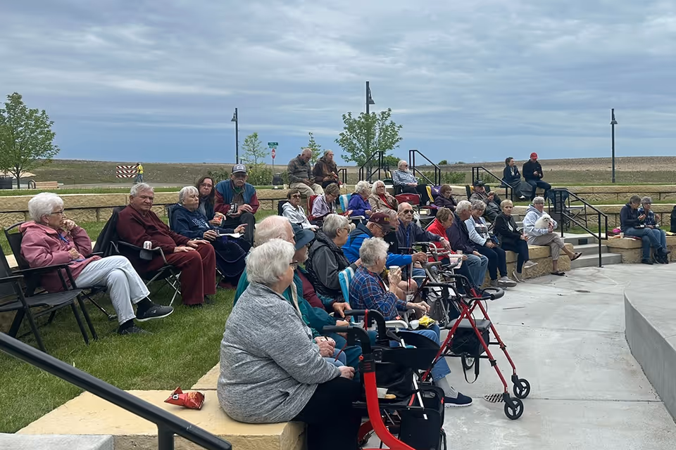 A group of elderly people sitting outdoors on benches and chairs arranged in a semi-circle, some using walkers, under a cloudy sky with a grassy area and trees in the background.