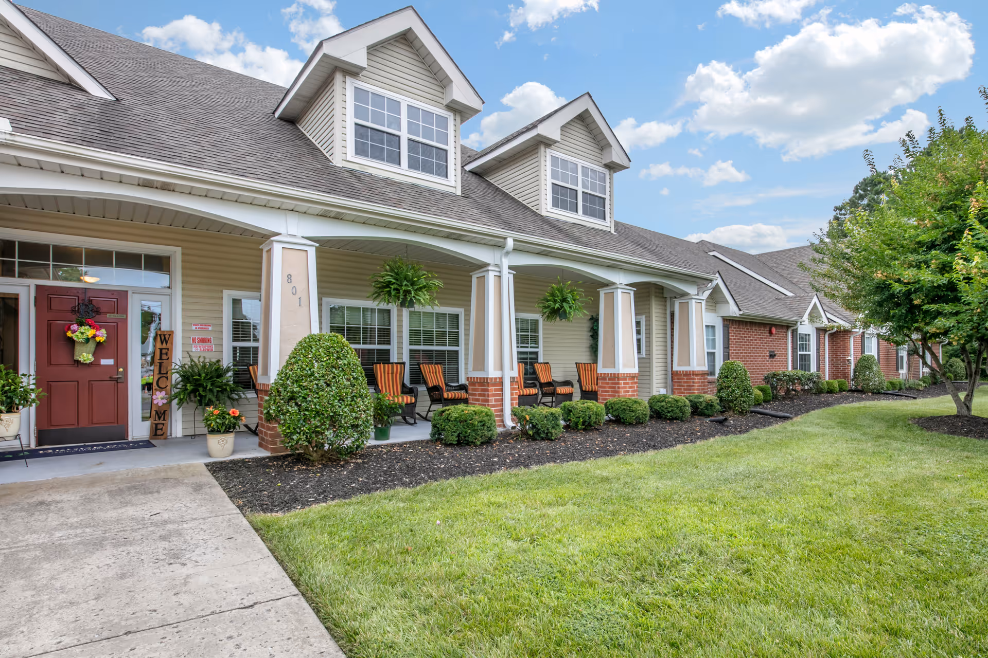Front exterior of a single-story senior living building with a covered porch, seating, landscaping, and a red entry door.