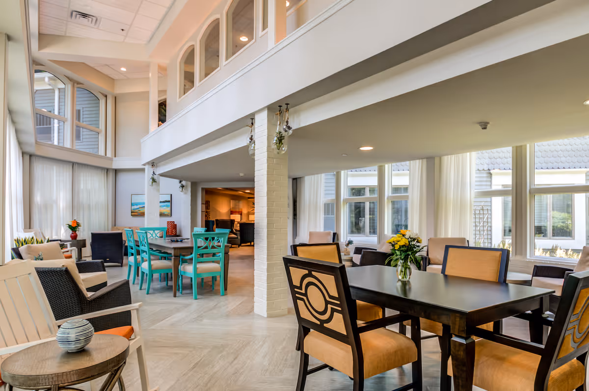 A bright and spacious common area in a senior living facility with large windows letting in natural light. The room features multiple seating arrangements including a black table with beige cushioned chairs, a round table with turquoise chairs, and various armchairs. There are flowers on the tables and artwork on the walls, creating a welcoming atmosphere.