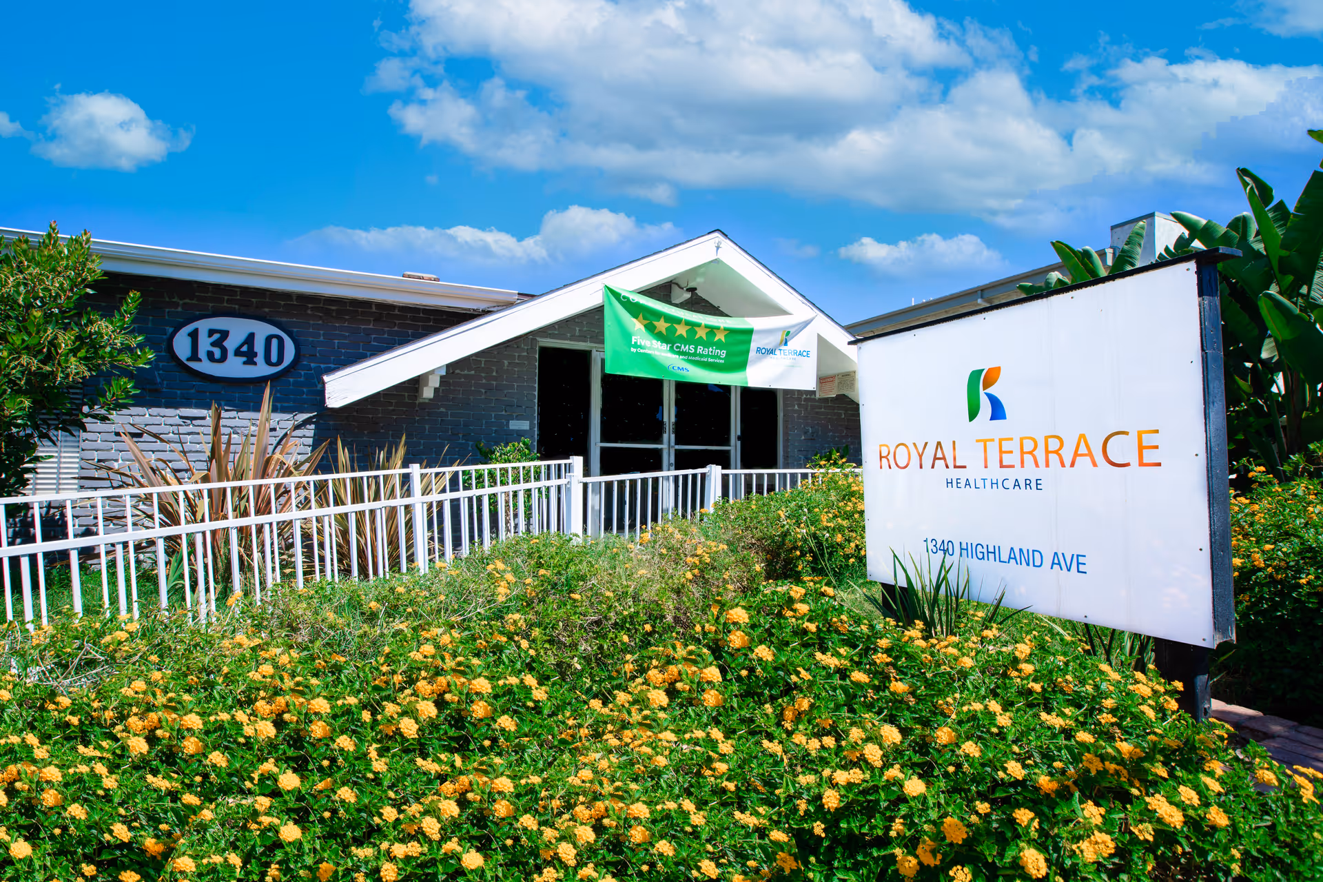 Front entrance of Royal Terrace Healthcare with its sign and address number 1340 surrounded by flowering landscaping under a blue sky.