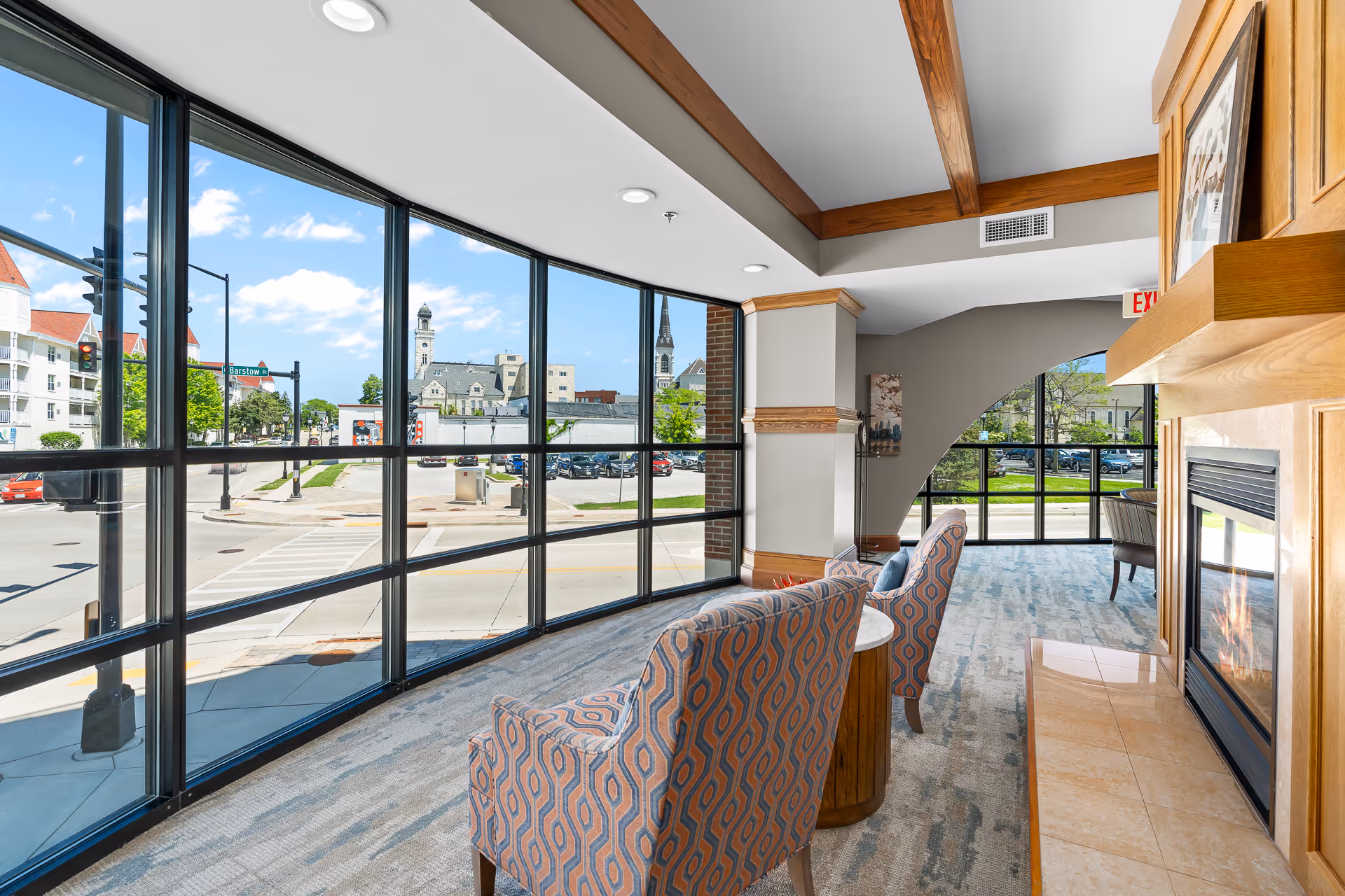 A bright and spacious lounge area with large floor-to-ceiling windows offering a view of the street outside. The room features two patterned armchairs facing a modern fireplace with a wooden mantle. The ceiling has wooden beams, and the carpet is a light, neutral color. Outside, buildings and a street intersection are visible under a blue sky with some clouds.