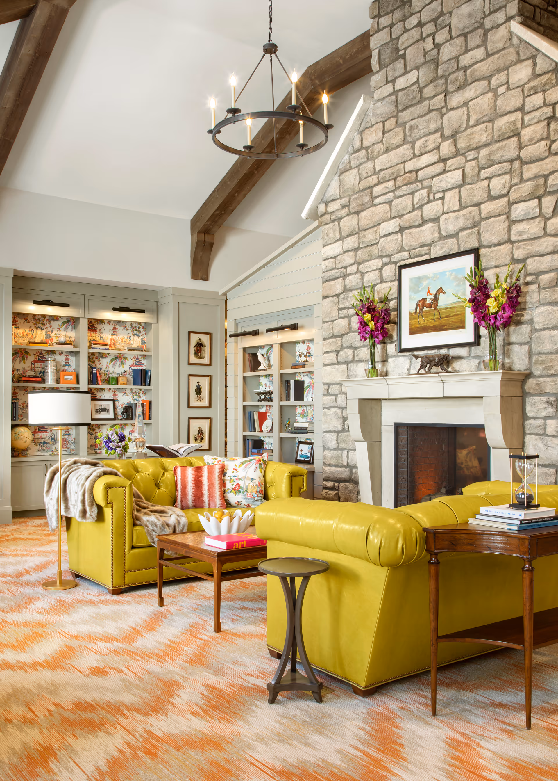 A cozy living room with two bright yellow leather sofas facing each other, a wooden coffee table with books and decorative items, a stone fireplace with a painting of a horse and rider above it, and two vases of purple flowers on the mantel. The room features exposed wooden beams on a high ceiling, built-in bookshelves with colorful wallpaper backing, and a patterned orange and beige carpet.