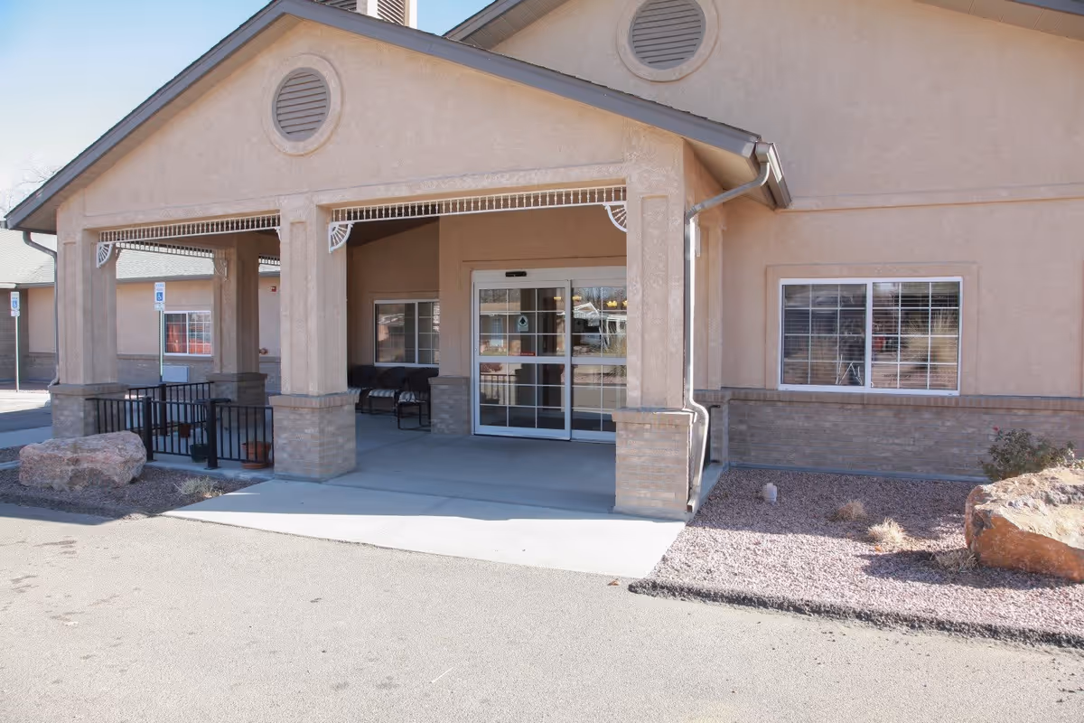 Entrance of a beige stucco building with a covered porch supported by columns, automatic sliding glass doors, and windows with white blinds. There are rocks and sparse landscaping with gravel around the building.