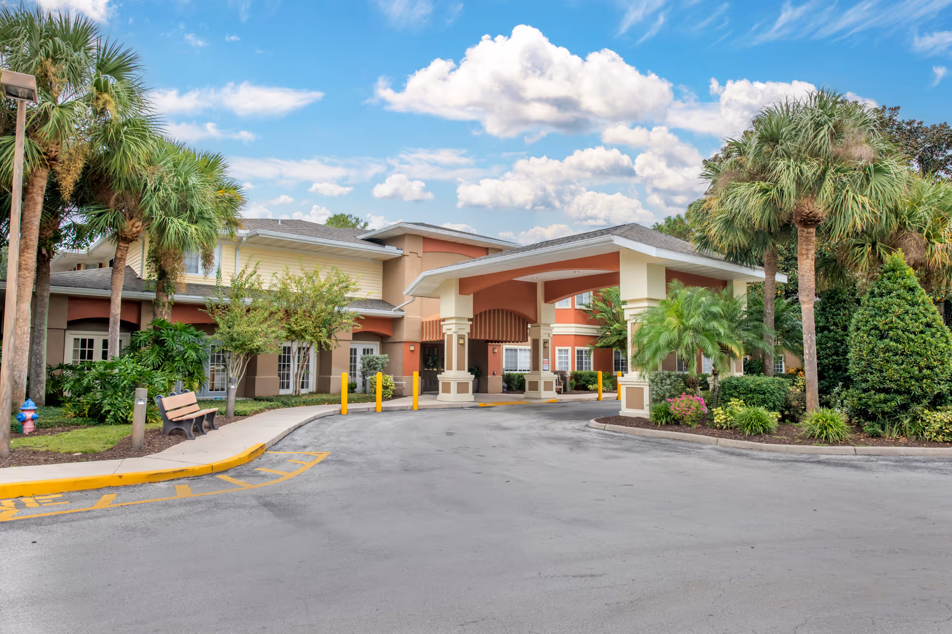 Front exterior view of Brookdale Altamonte Springs facility showing a covered entrance with columns, surrounded by palm trees and landscaped greenery under a partly cloudy blue sky.