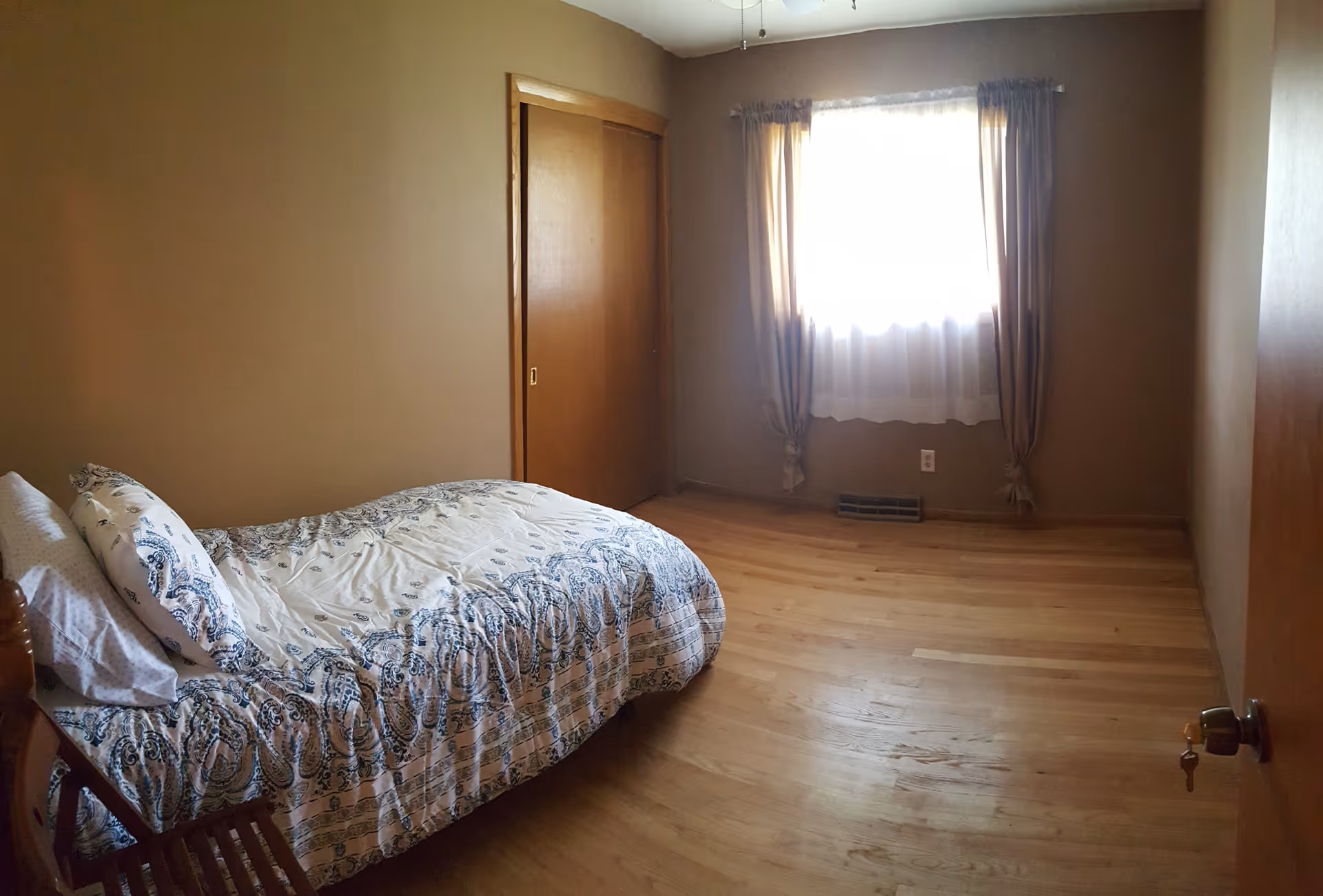 A simple bedroom with a single bed covered in a white and blue patterned bedspread. The room has wooden floors, beige walls, a window with sheer and brown curtains, and a wooden closet door. A wooden chair is partially visible next to the bed.