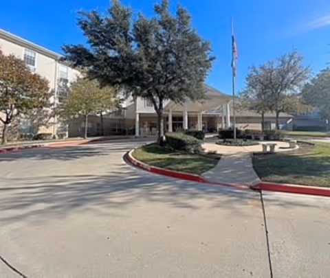 Front entrance of a senior living building with a circular driveway, flagpole, trees, and benches.