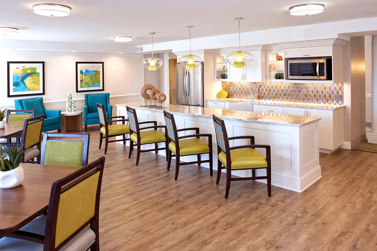 A bright and modern communal kitchen and dining area in an assisted living facility. The space features a long white kitchen island with a granite countertop and four chairs with green cushions. Behind the island is a kitchen area with white cabinets, a stainless steel refrigerator, a microwave, and a hexagonal tile backsplash. To the left, there are dining tables with chairs that have green backs and seats, and two teal armchairs with a small round table between them. The walls are decorated with colorful abstract paintings, and the floor is a light wood finish.