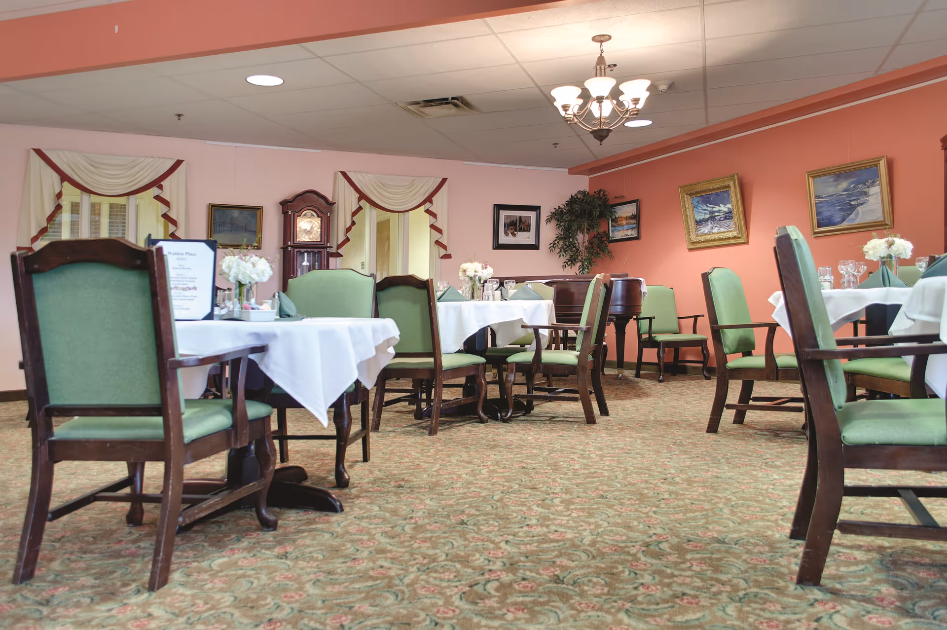 A dining room with several tables covered in white tablecloths and set with glassware and floral centerpieces. The chairs have green upholstery and dark wooden frames. The room has a patterned carpet, peach-colored walls with framed paintings, a grandfather clock, and a chandelier hanging from the ceiling.