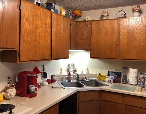 A kitchen corner with wooden cabinets, a double sink, and various kitchen items including a red coffee maker, bananas, a blender, paper towels, and decorative figurines on the countertop and above the cabinets.