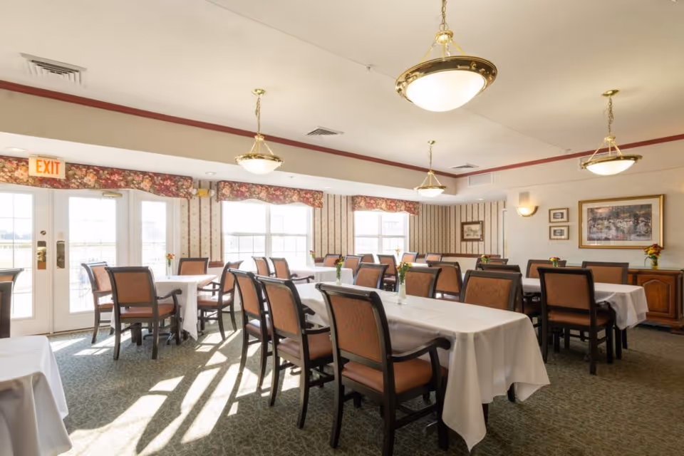 A bright dining room with multiple tables covered in white tablecloths and surrounded by brown cushioned chairs. The room has large windows letting in natural light, floral valances, patterned wallpaper, and several ceiling light fixtures. There are small flower vases on the tables and framed artwork on the walls.
