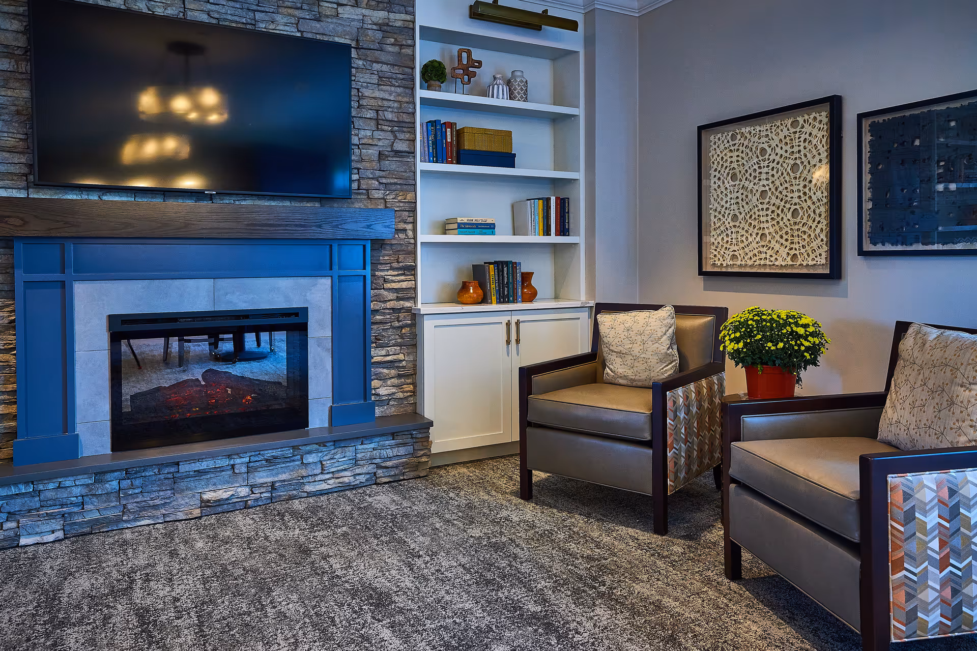Cozy living room area with a stone fireplace and a mounted flat-screen TV above it. Two cushioned armchairs with patterned sides are positioned near a small table holding a potted yellow flower. A built-in white bookshelf with books and decorative items is next to the fireplace. Two framed abstract artworks hang on the wall behind the chairs.