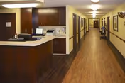Interior view of a healthcare facility hallway with wood flooring and beige walls. On the left side, there is a reception desk with a computer and office supplies. The hallway has several closed doors on both sides and framed pictures hanging on the walls. The area is well-lit with ceiling lights.