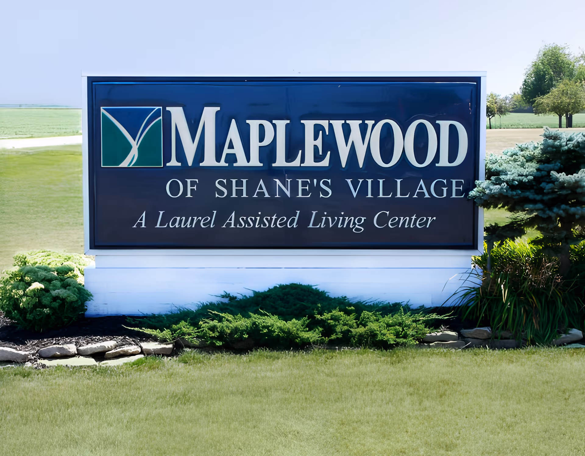 Outdoor sign for Maplewood of Shane's Village, a Laurel Assisted Living Center, surrounded by grass, shrubs, and trees under a clear sky.