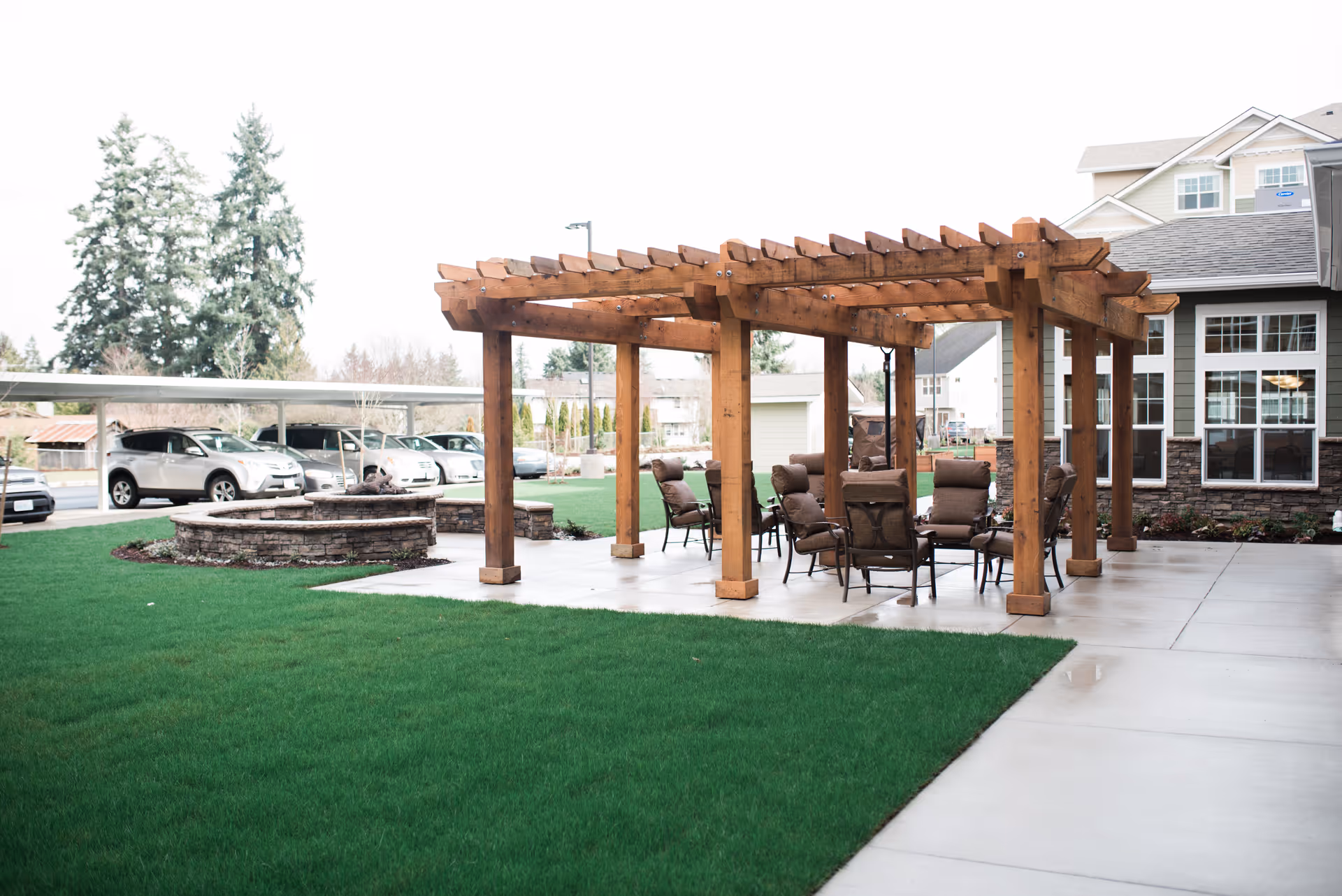 Outdoor patio area at Julian Estates Gracious Retirement Living featuring a wooden pergola with cushioned chairs arranged underneath. There is a circular stone fire pit nearby, surrounded by green grass and concrete walkways. Several parked cars and trees are visible in the background.