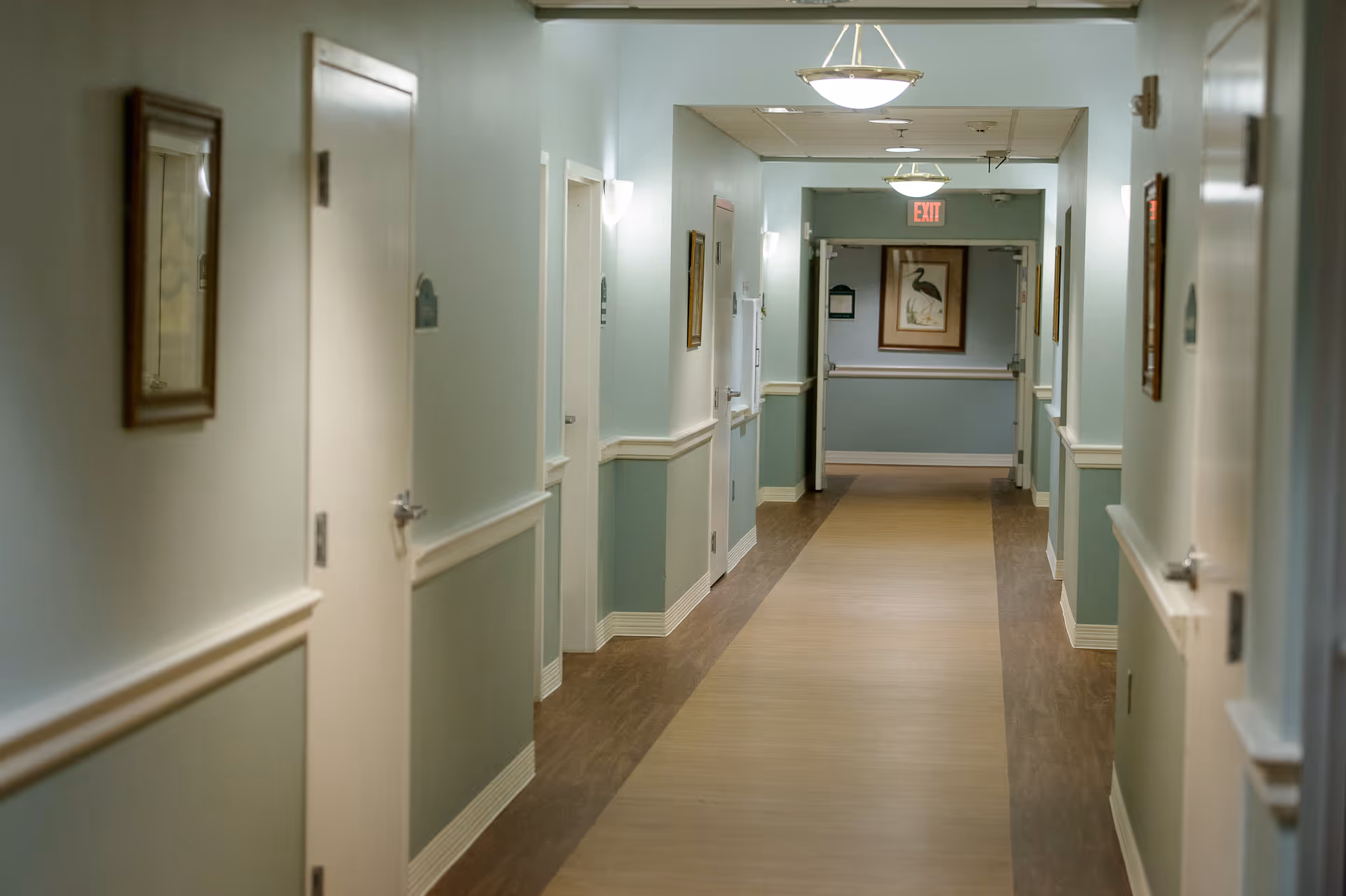 A well-lit hallway in a senior living facility with light blue walls, white doors on both sides, framed artwork, and ceiling lights. The floor has a wood-like border with a lighter center path, and an exit sign is visible at the end of the corridor.