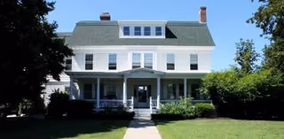 A white, multi-story senior living building with a front porch and a well-kept lawn.