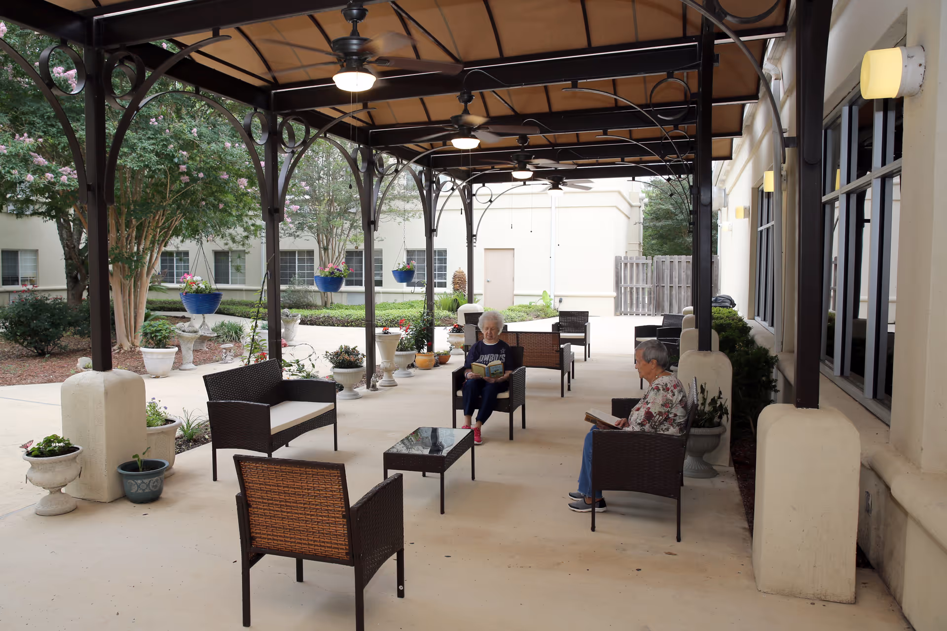 Two elderly women sitting on outdoor wicker chairs under a covered patio with ceiling fans, reading books. The patio is decorated with potted plants and hanging flower pots, with a garden and building visible in the background.