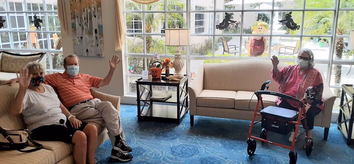 Three elderly people wearing face masks sitting on sofas in a bright living room with large windows showing an outdoor garden. Two people sit on a beige sofa on the left, and one person with a red walker sits on a beige sofa on the right. They are waving at the camera. The room has a blue patterned carpet, a side table with a lamp, and Halloween-themed decorations on the windows.