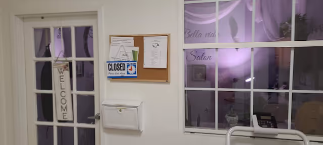 Interior view of a hallway or entrance area with a door on the left that has a hanging sign reading 'WELCOME'. Next to the door is a bulletin board with various notices, including a 'CLOSED' sign. To the right, there is a window with the words 'Bella vida Salon' visible through it, showing a softly lit room with purple lighting and some salon equipment.
