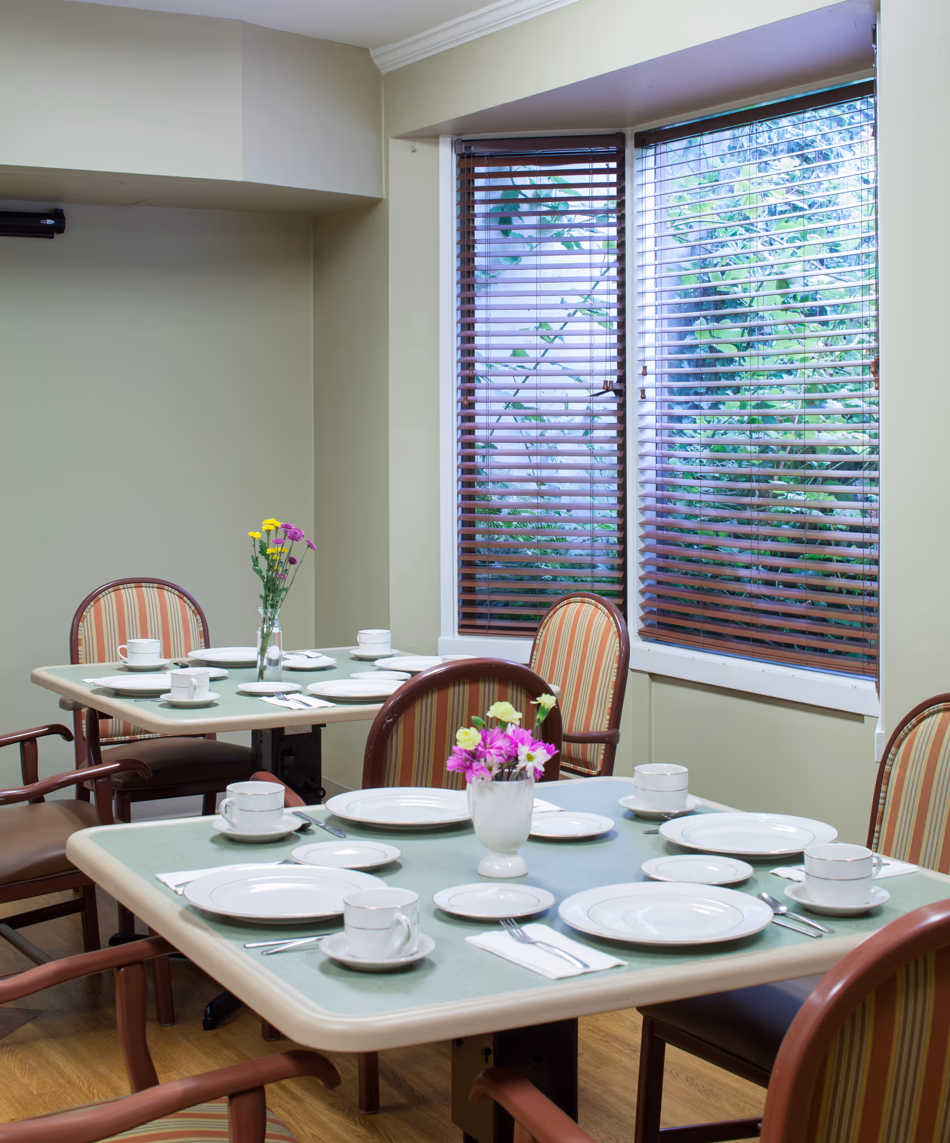 A dining room with two tables set for a meal, each with white plates, cups, and silverware. The tables have green placemats and small vases with colorful flowers. There are striped upholstered chairs around the tables and a window with wooden blinds showing greenery outside.