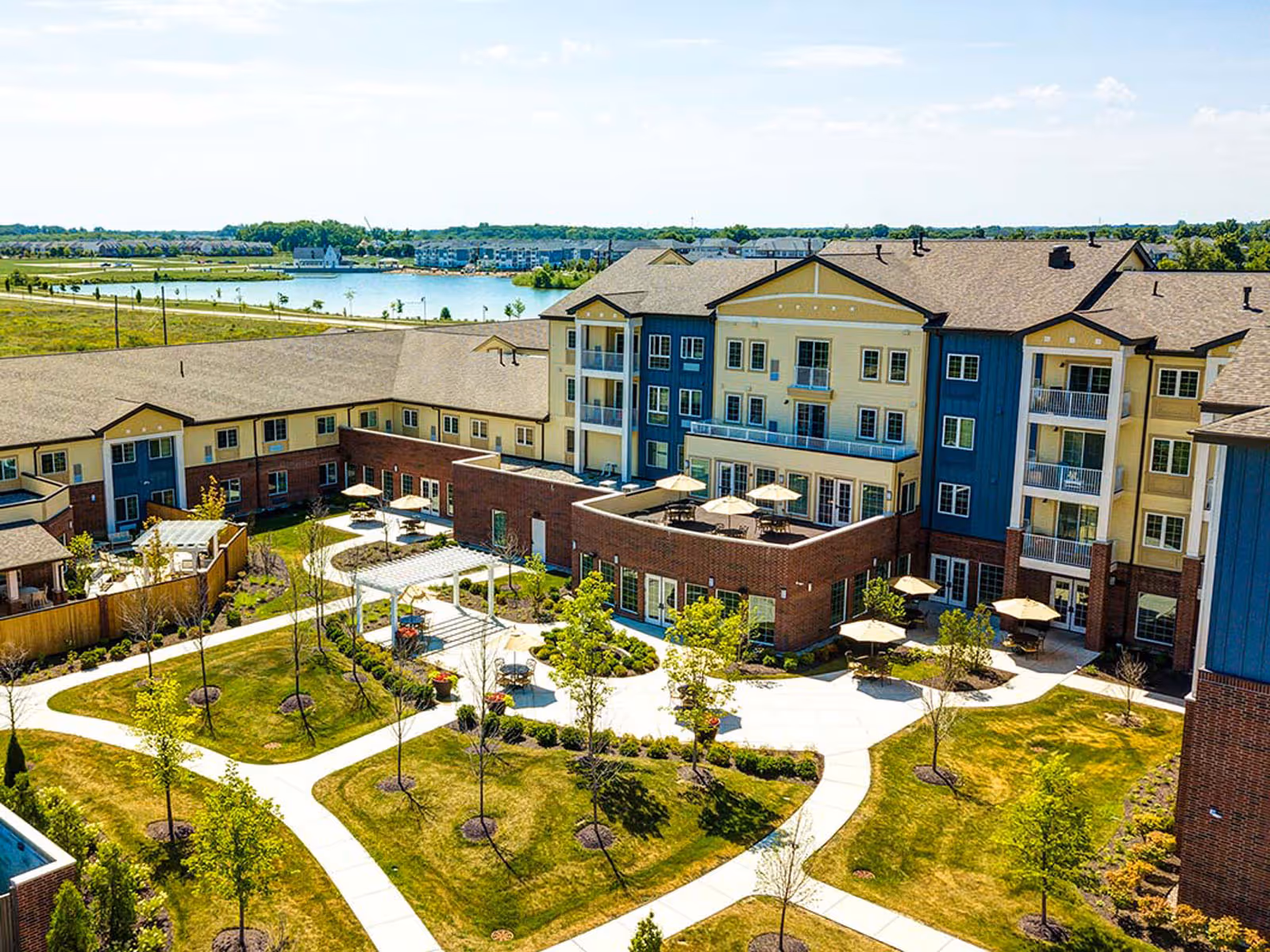 Aerial view of Independence Village Fishers East showing a multi-story senior living building with balconies, surrounded by landscaped gardens, walking paths, and outdoor seating areas with umbrellas. In the background, there is a lake and additional residential buildings under a partly cloudy sky.