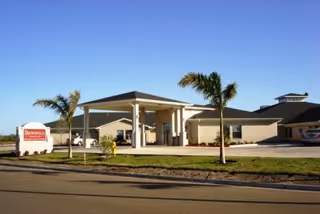Exterior view of Brownsville Nursing and Rehabilitation Center showing a single-story building with a covered entrance, palm trees, and a clear blue sky.