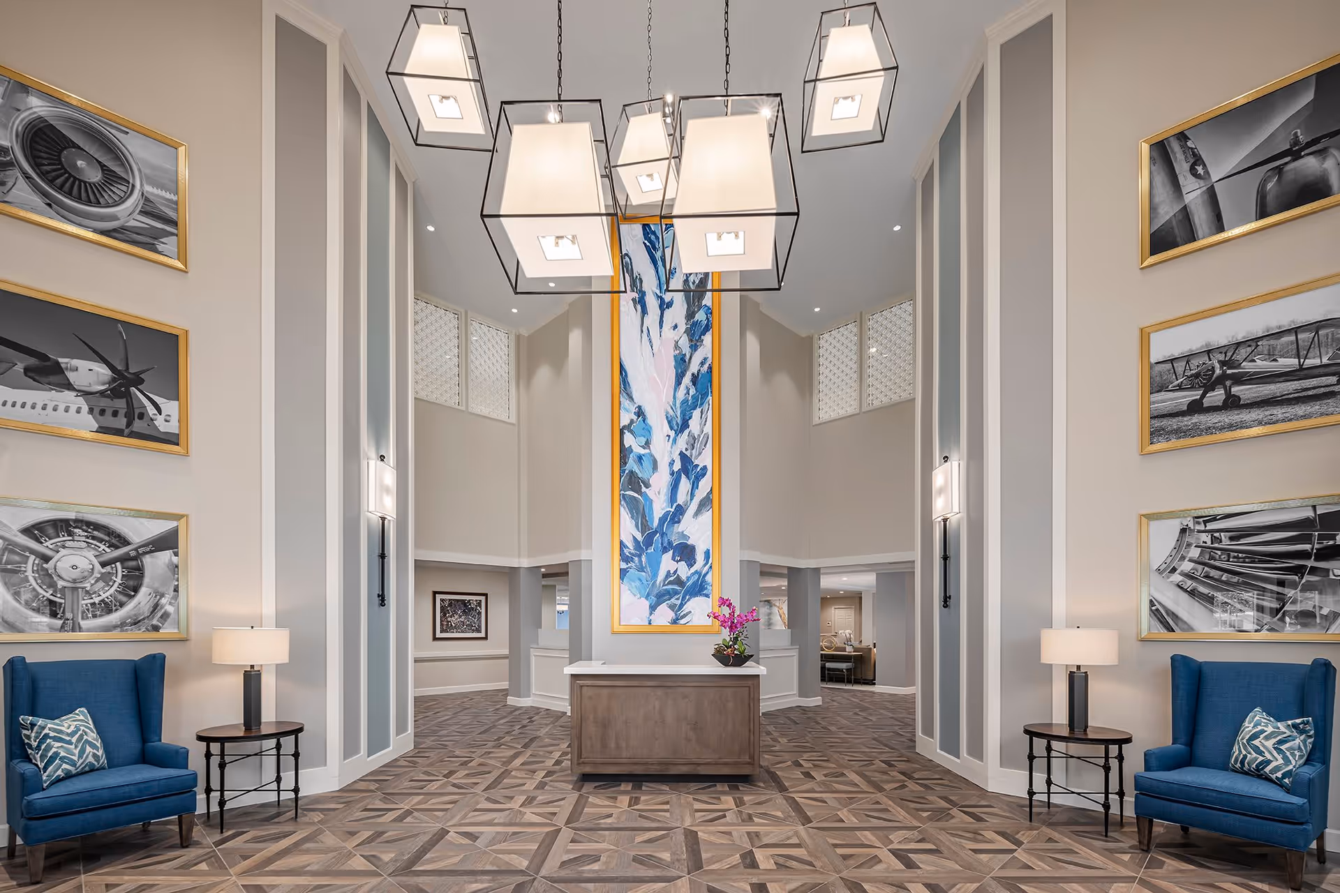 Spacious and elegant lobby area with high ceilings, modern hanging light fixtures, and a central wooden reception desk adorned with a flower arrangement. The walls feature large framed black and white photographs of airplane parts and a tall colorful abstract painting in the center. Two blue upholstered armchairs with patterned pillows flank small round tables with lamps on either side of the room. The floor has a geometric wood pattern.