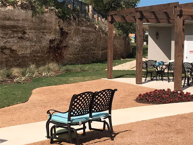 Outdoor seating area with two black metal benches with light blue cushions on a gravel path, a wooden pergola with a table and chairs underneath, surrounded by grass, plants, and a rocky cliff wall in the background.