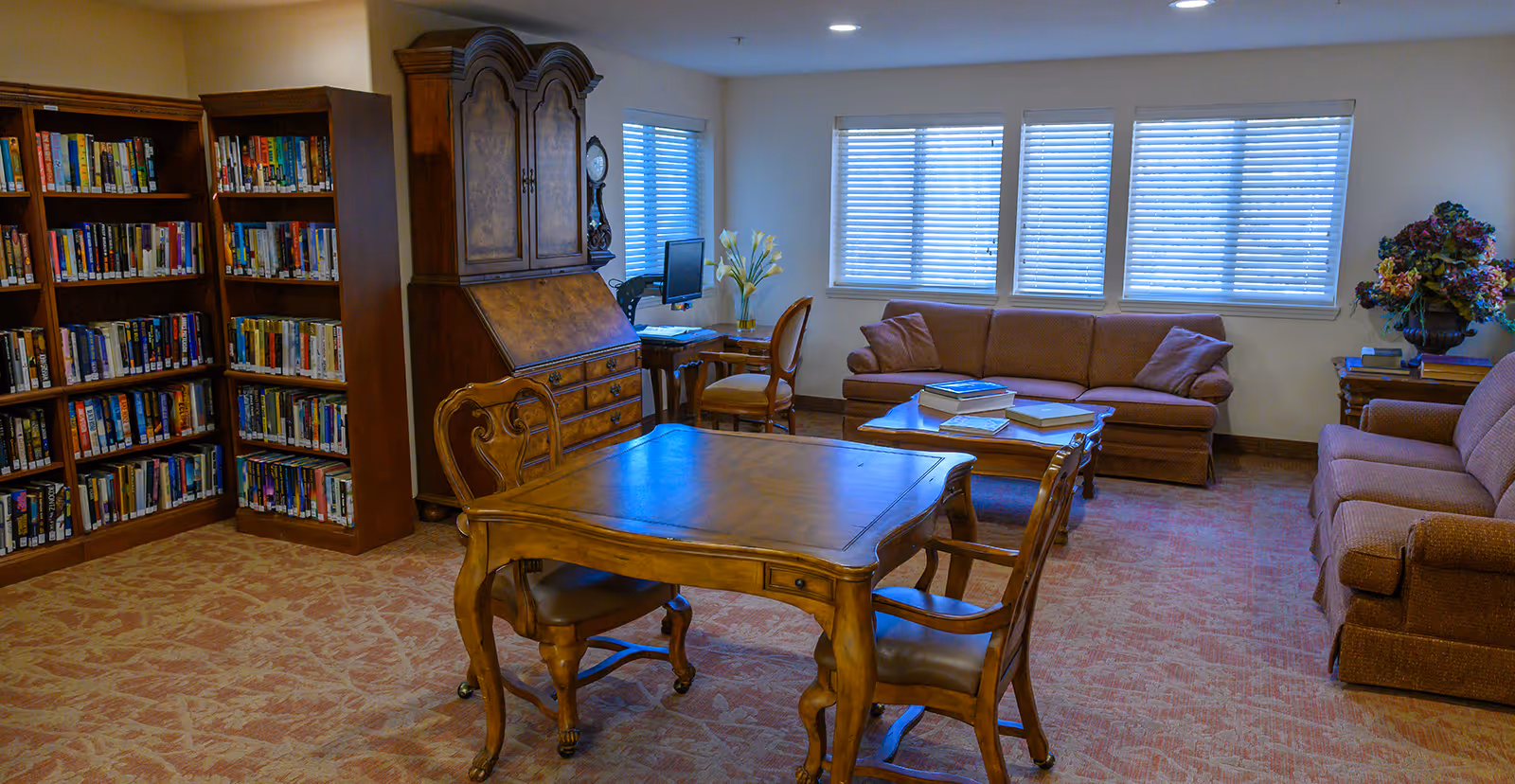 A cozy living room area with two brown upholstered sofas, a wooden coffee table with books on it, a wooden table with two chairs in the foreground, bookshelves filled with books on the left, a wooden cabinet, a small desk with a computer, and windows with blinds letting in natural light.