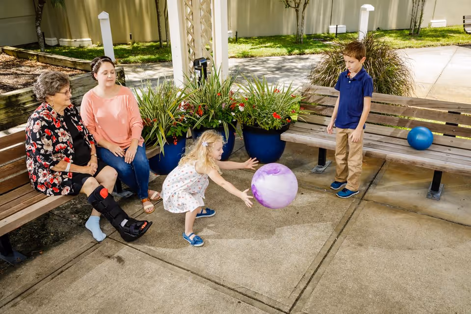 An elderly woman with a leg brace and a younger woman sit on a wooden bench outdoors near potted plants. A young girl in a white dress with polka dots is playing with a purple ball, while a young boy in a blue shirt and khaki pants stands nearby watching. There is another blue ball on a separate bench in the background.