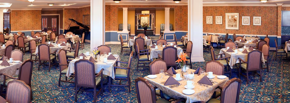Spacious formal dining room in an assisted living facility with multiple set tables and chairs, decorative carpeting, columns, and a grand piano in the back.