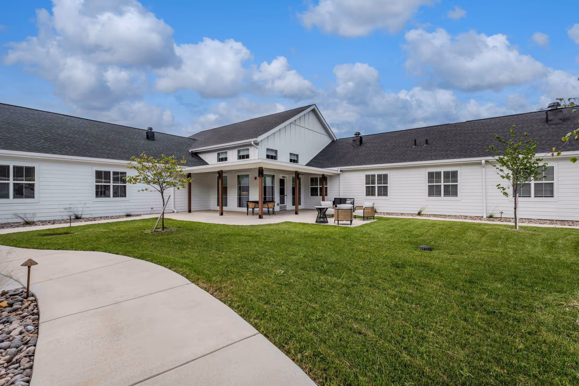Outdoor courtyard with a green lawn, patio seating, and a white single-story building under a partly cloudy sky.