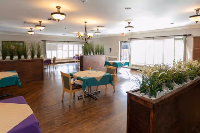 Spacious dining room with small tables covered in green and purple tablecloths, wooden chairs, large planter dividers, and windows letting in natural light.