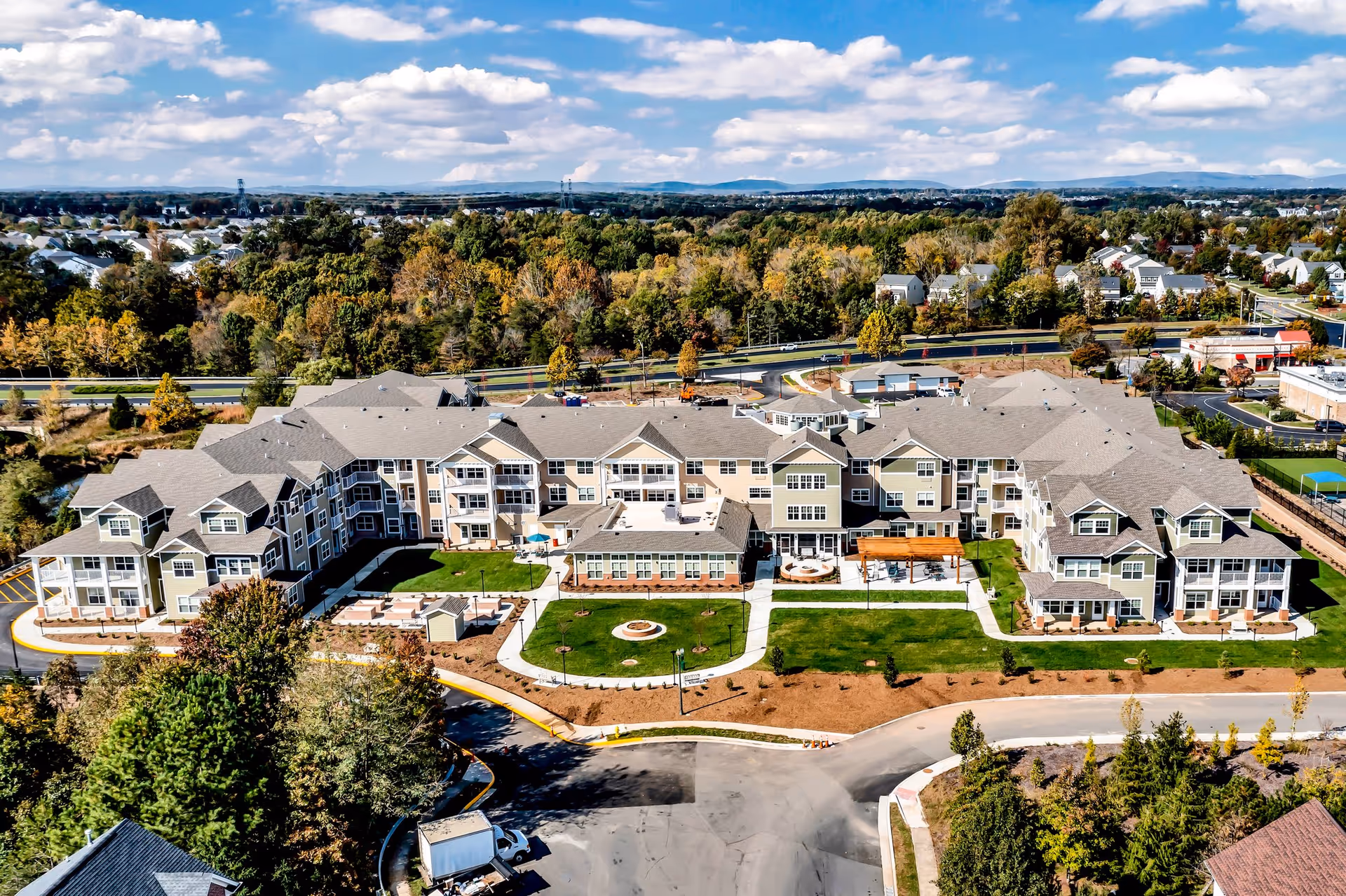 Aerial view of Capitol Ridge Gracious Retirement Living facility showing a large, multi-wing building with beige and green exterior walls and gray roofs. The building is surrounded by landscaped lawns, pathways, and trees with a backdrop of a suburban neighborhood and distant hills under a partly cloudy sky.