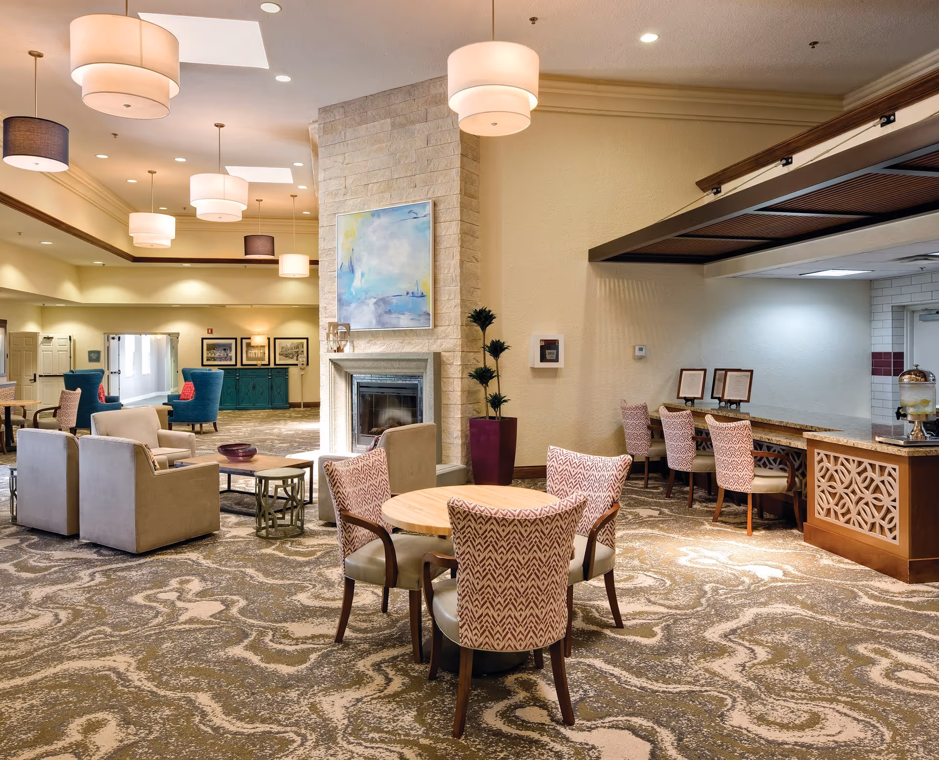 A spacious and well-lit common area in a senior living facility featuring a round wooden table with four patterned chairs in the foreground. Behind it, there is a stone fireplace with a colorful abstract painting above it. To the right, there is a counter with four chairs and framed menus or notices. The room has a patterned carpet, multiple ceiling lights, and additional seating areas with armchairs and small tables in the background.