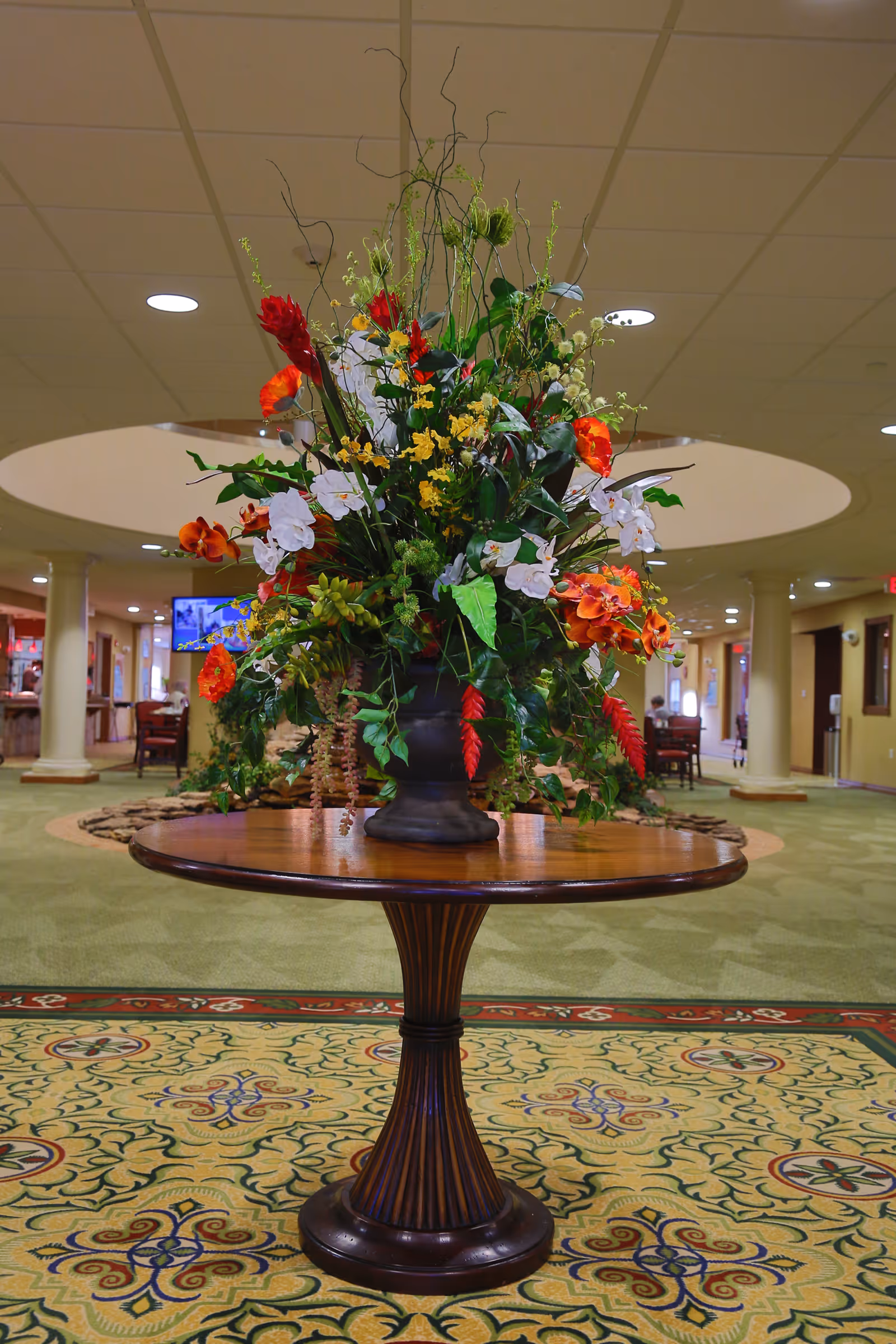 A large floral arrangement with red, orange, yellow, and white flowers and green leaves sits on a round wooden table in the center of a carpeted room with columns and recessed lighting.