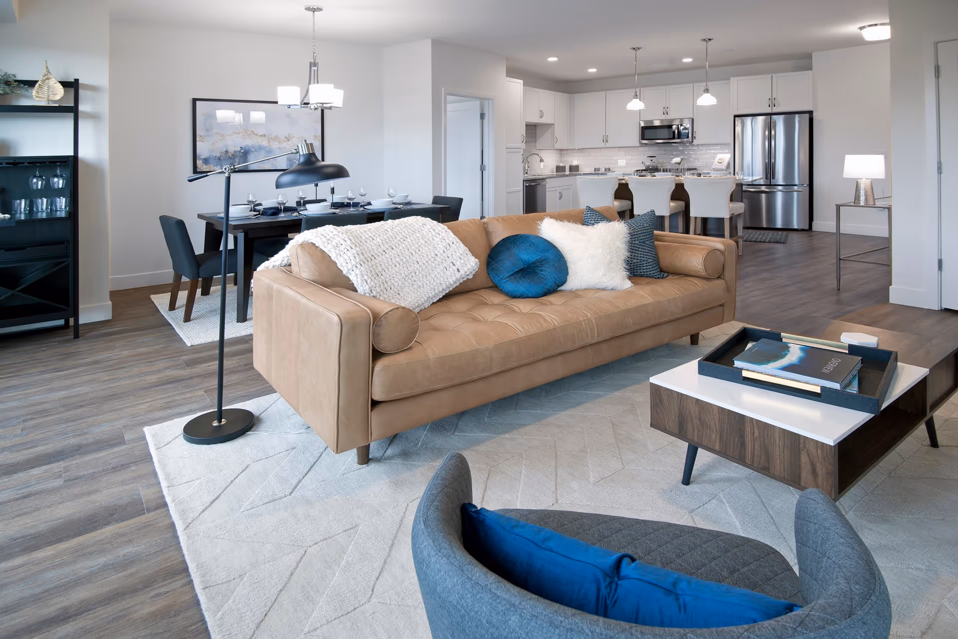 Modern open-concept living space featuring a tan leather sofa with blue and white pillows, a gray armchair with a blue cushion, a wooden coffee table with books and a tray, a black floor lamp, a dining area with a dark wood table set for six, and a kitchen with white cabinets, stainless steel appliances, and a breakfast bar with four beige stools.